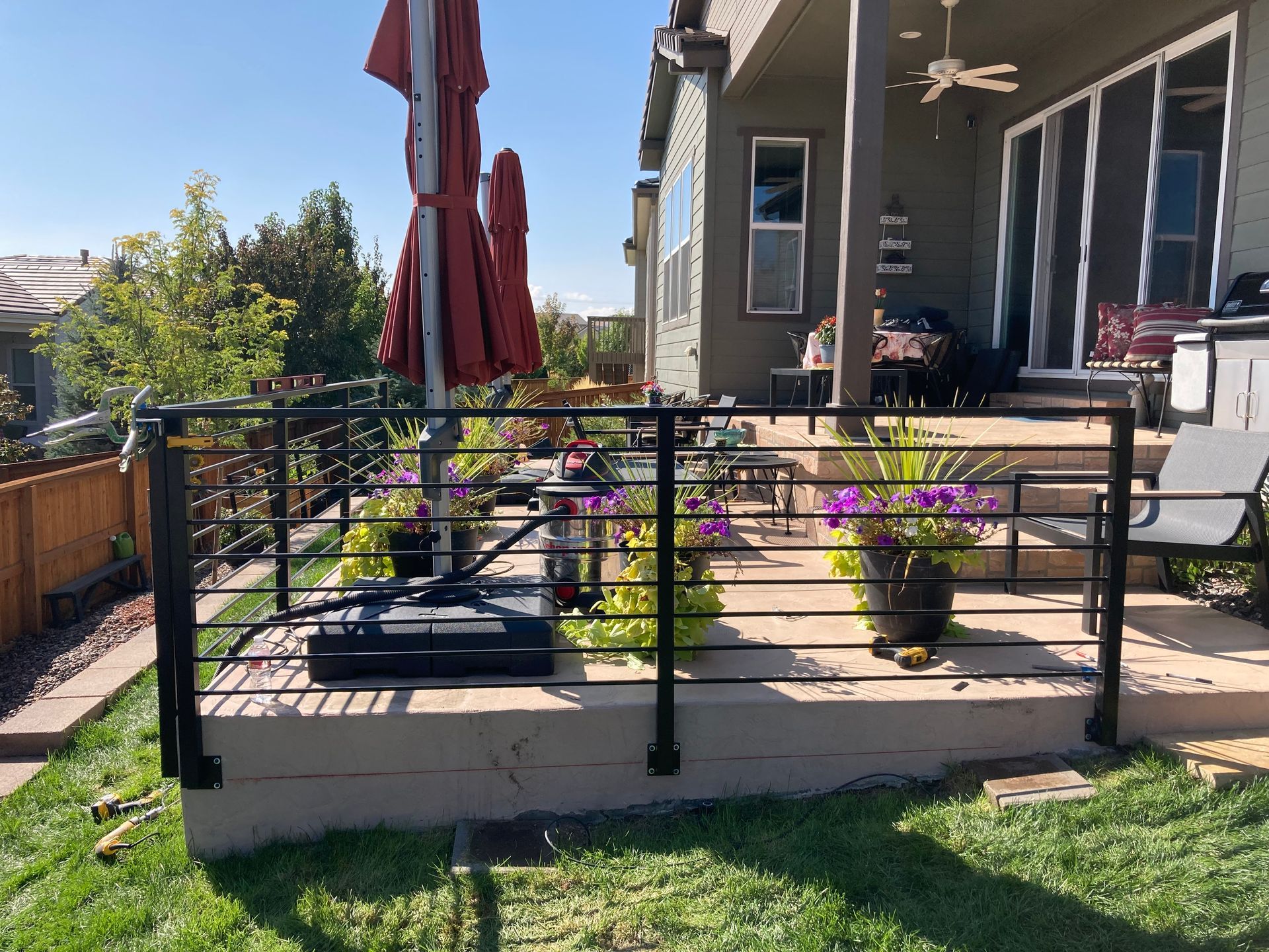 Black metal railing on a patio with potted flowers, an umbrella, and a view of a house.