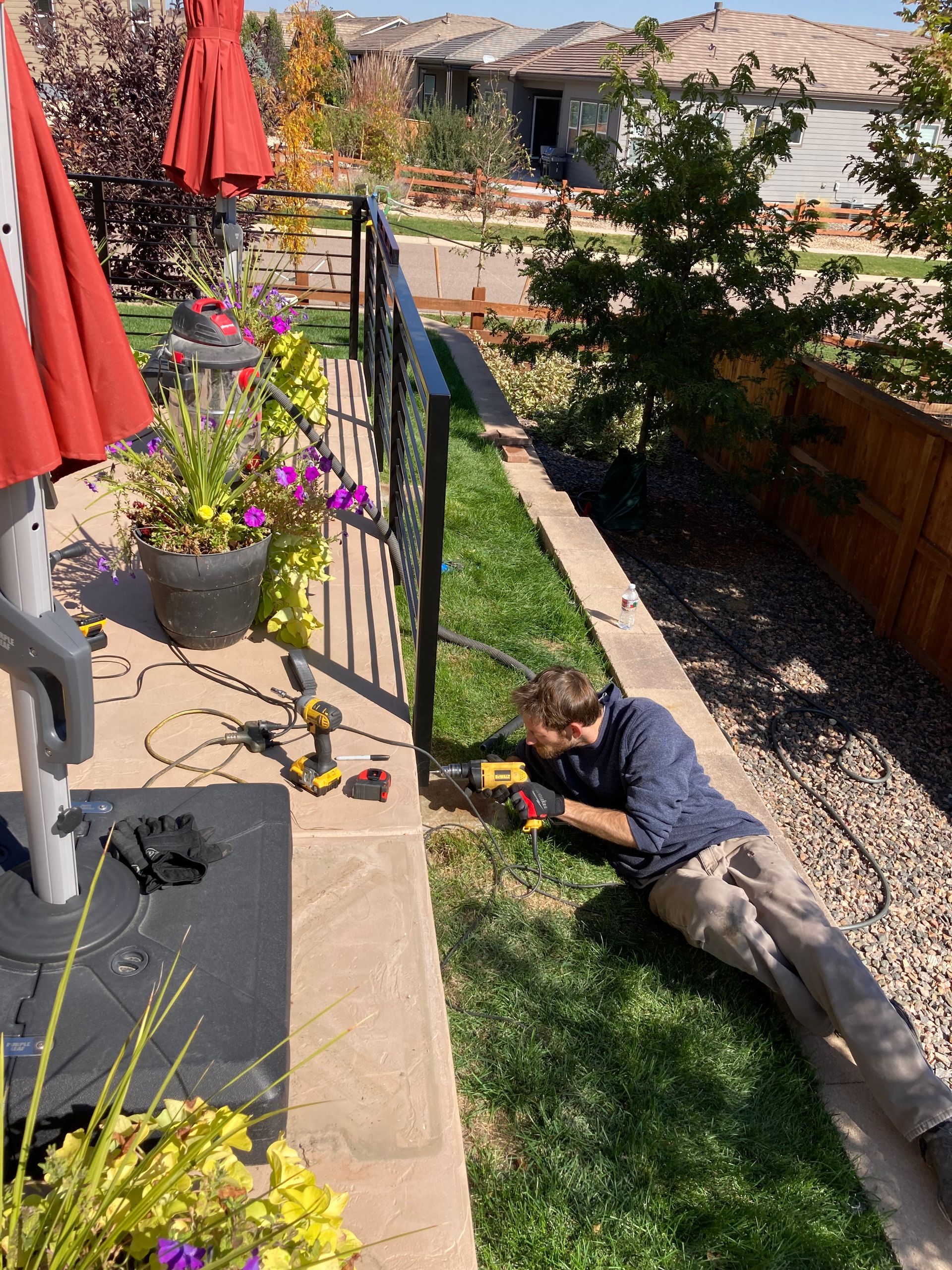 Man using a drill to install a black railing along a sidewalk. Outdoors, sunny day.