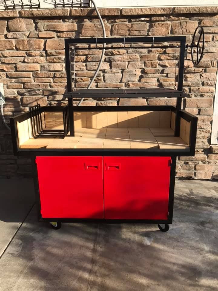 Red and black outdoor grill on wheels with brick backdrop. Features a rotisserie and storage cabinet.