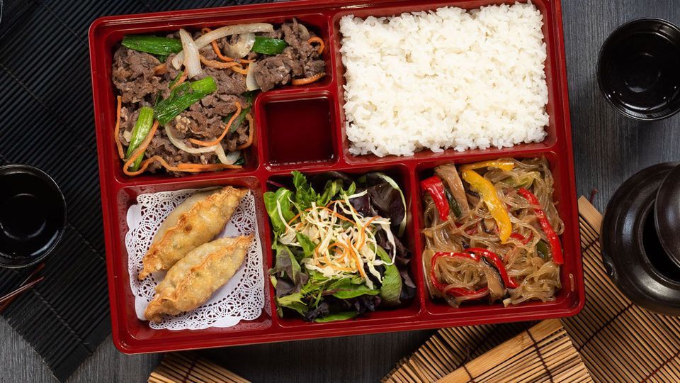 Red bento box with rice, grilled meat, dumplings, and vegetables on a dark table with tea cups