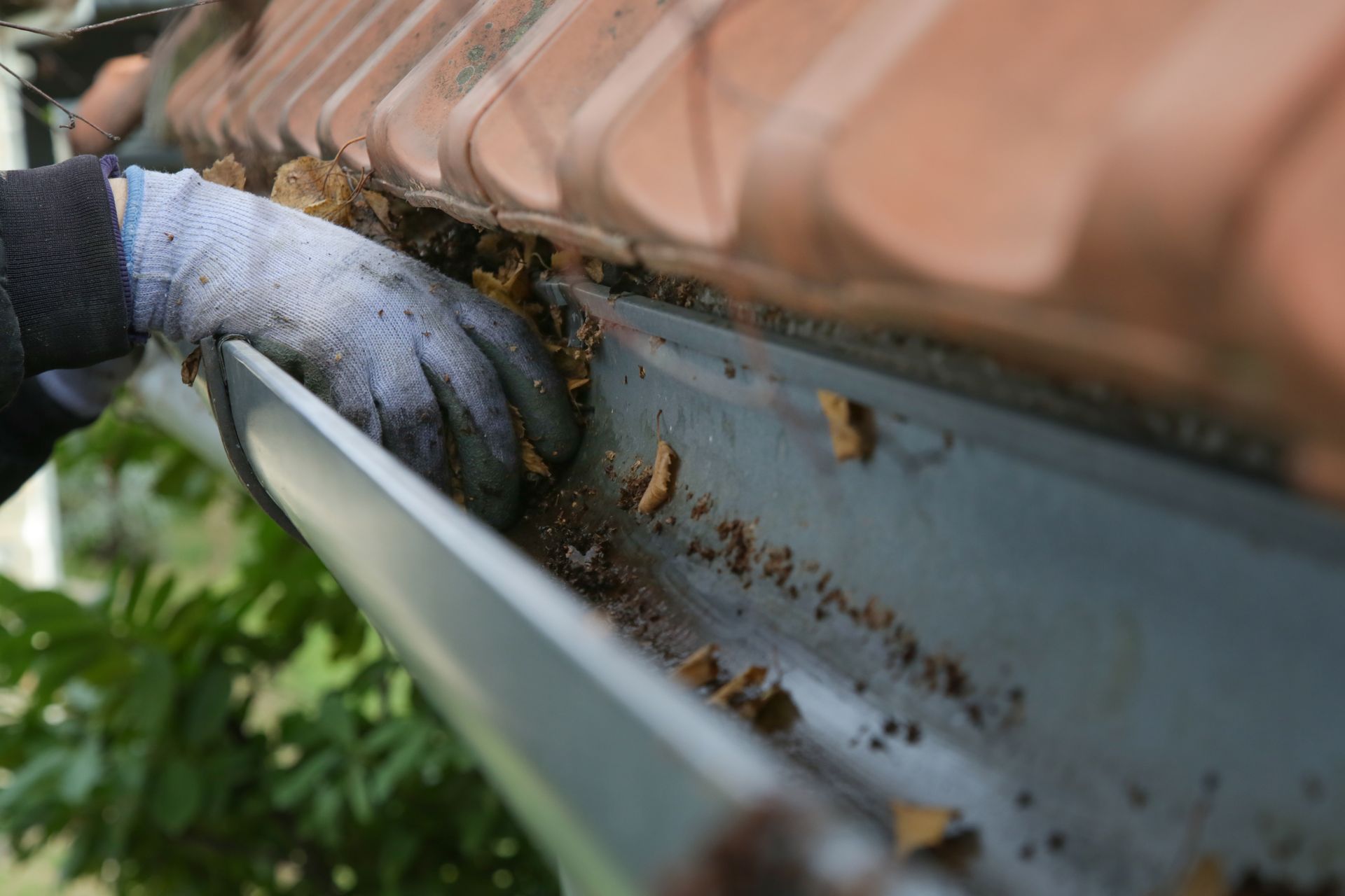 close-up of a gloved hand cleaning leaves and debris from a metal rain gutter beneath a terracotta 