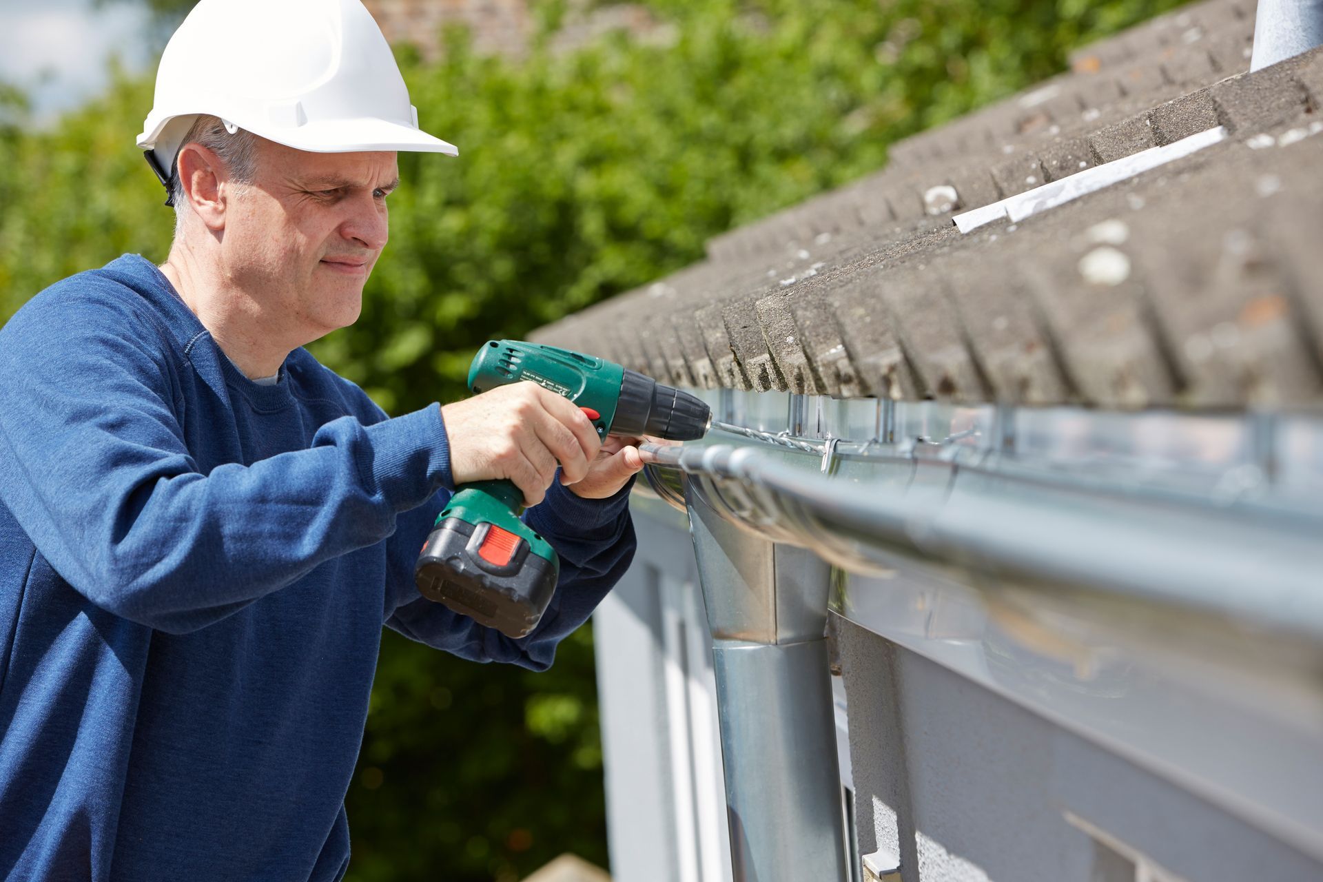 A worker in a helmet uses a drill to replace a roof gutter during home maintenance. A worker in a helmet uses a drill to replace a roof gutter during home maintenance.