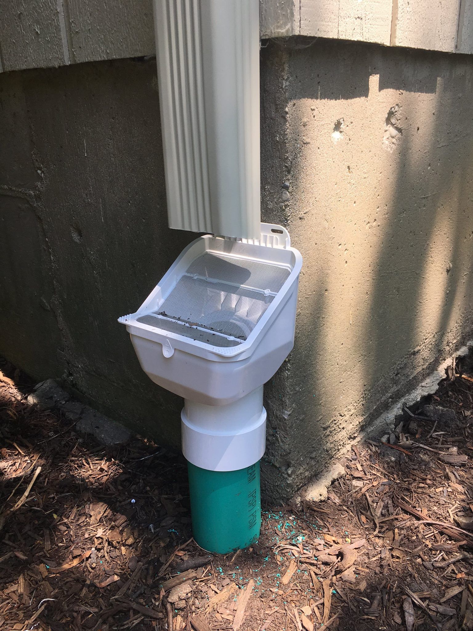 White downspout connected to a screened drain, extending to a green pipe, against a concrete wall.
