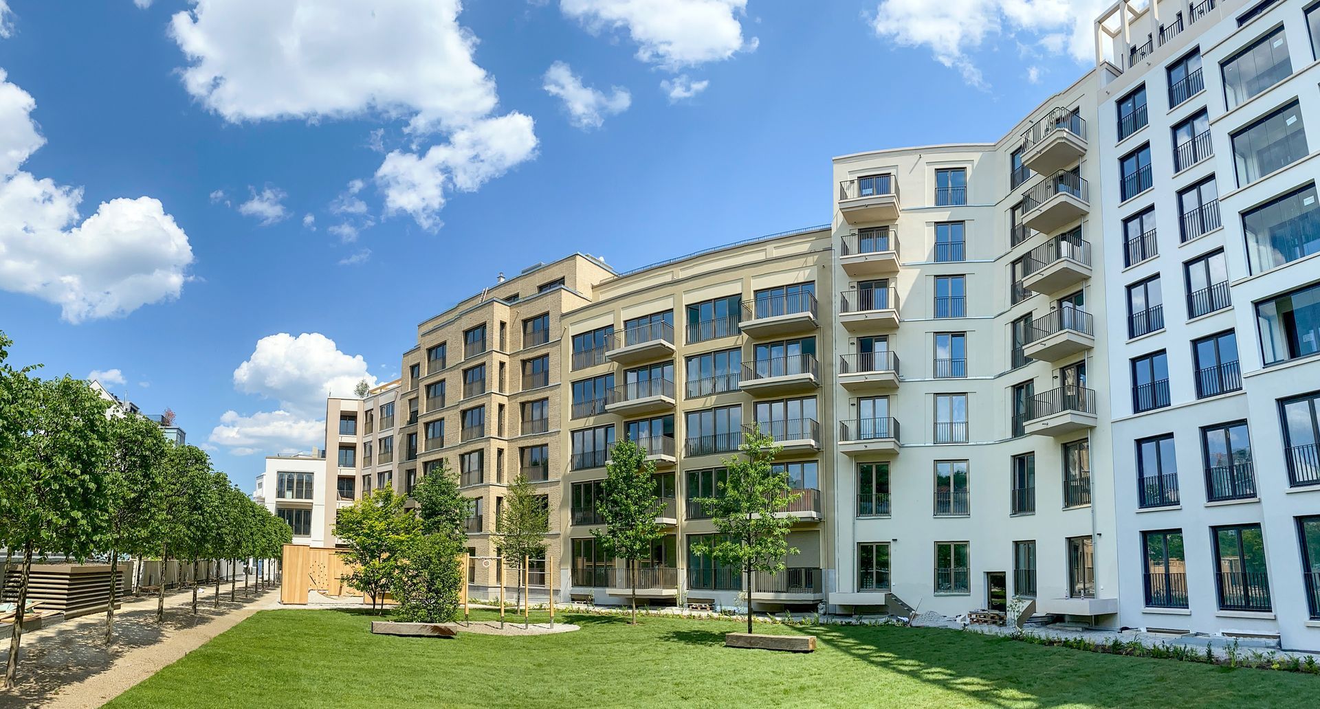 Modern apartment buildings with balconies against a blue sky, green lawn and trees in front.