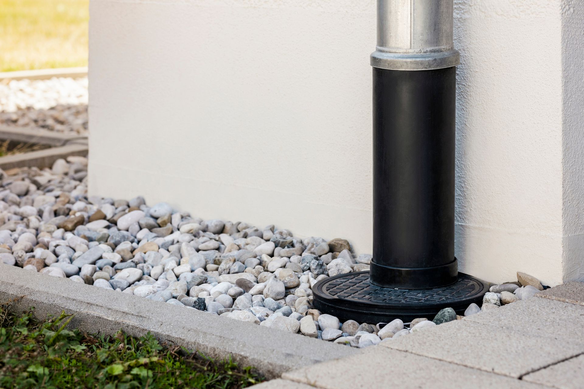 Downspout attached to a wall, surrounded by river rocks and bordering a concrete path.
