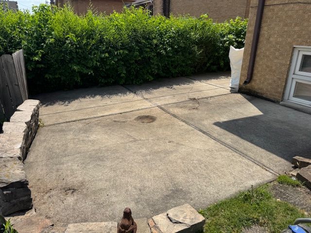 Concrete patio next to a building and fence, with a green hedge and small patch of grass.
