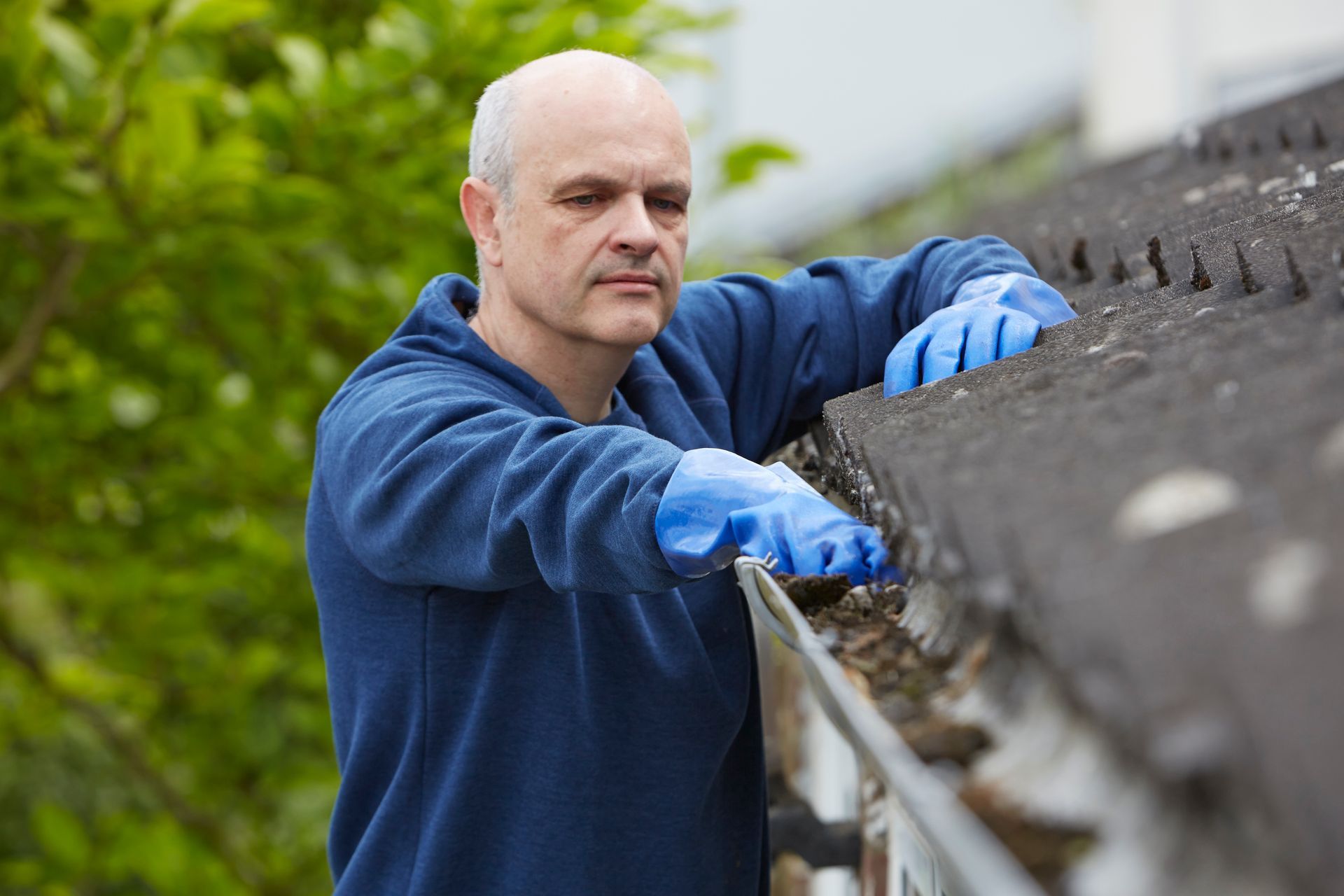 A man on a ladder, performing gutter cleaning service near trees under a clear blue sky.