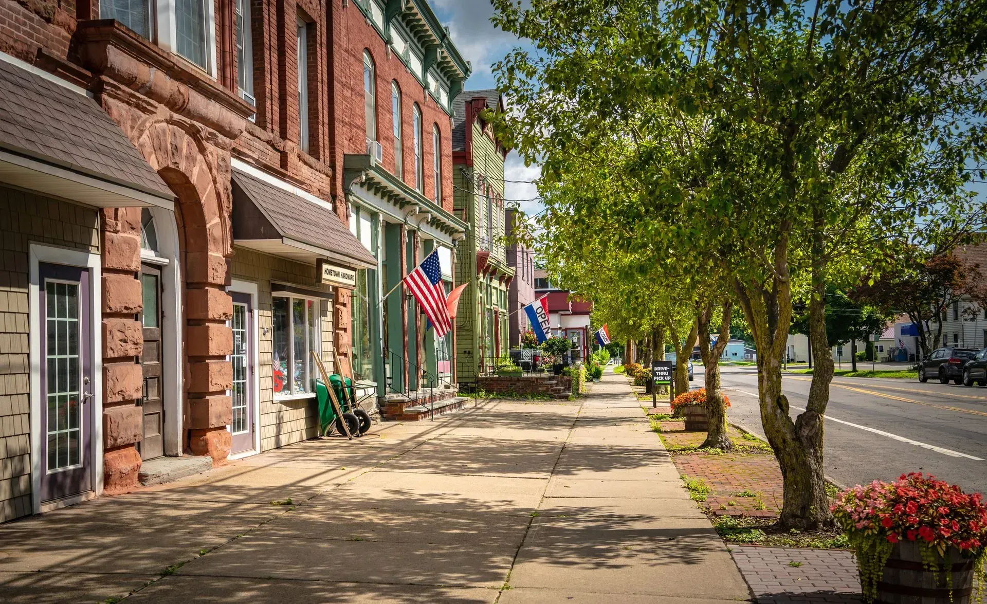 A sidewalk along a quiet main street lined with historic brick storefronts, American flags, and green leafy trees. A sidewalk along a quiet main street lined with historic brick storefronts, American flags, and green leafy trees.