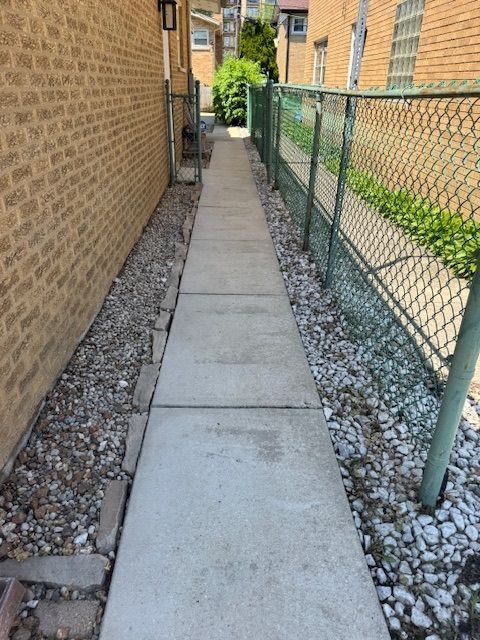 Narrow concrete walkway between a brick building and chain-link fence, with gravel on either side.