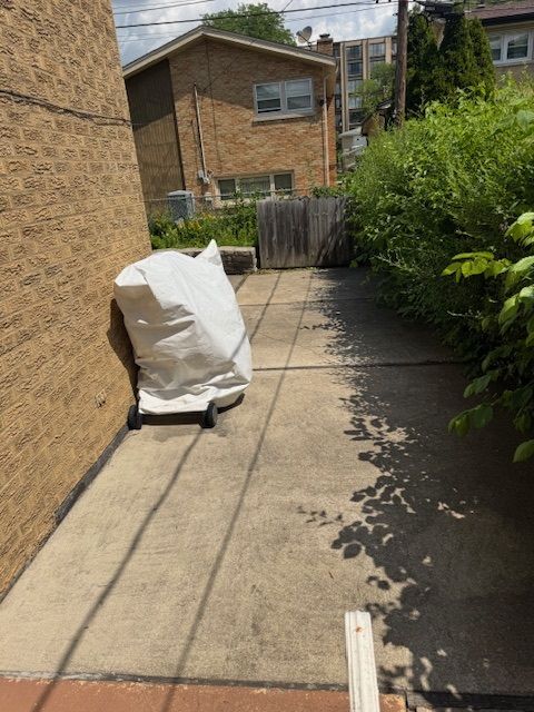 Concrete pathway between brick building and overgrown bushes; covered grill on the left.