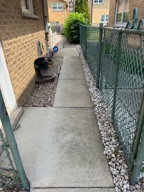Concrete pathway alongside a brick building, separated by a chain-link fence and gravel.