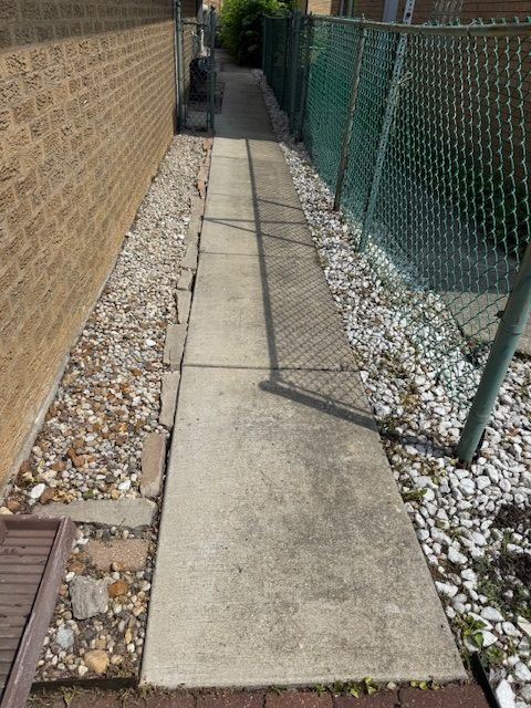 Concrete walkway between a brick wall and a chain-link fence, with gravel and small rocks on either side.