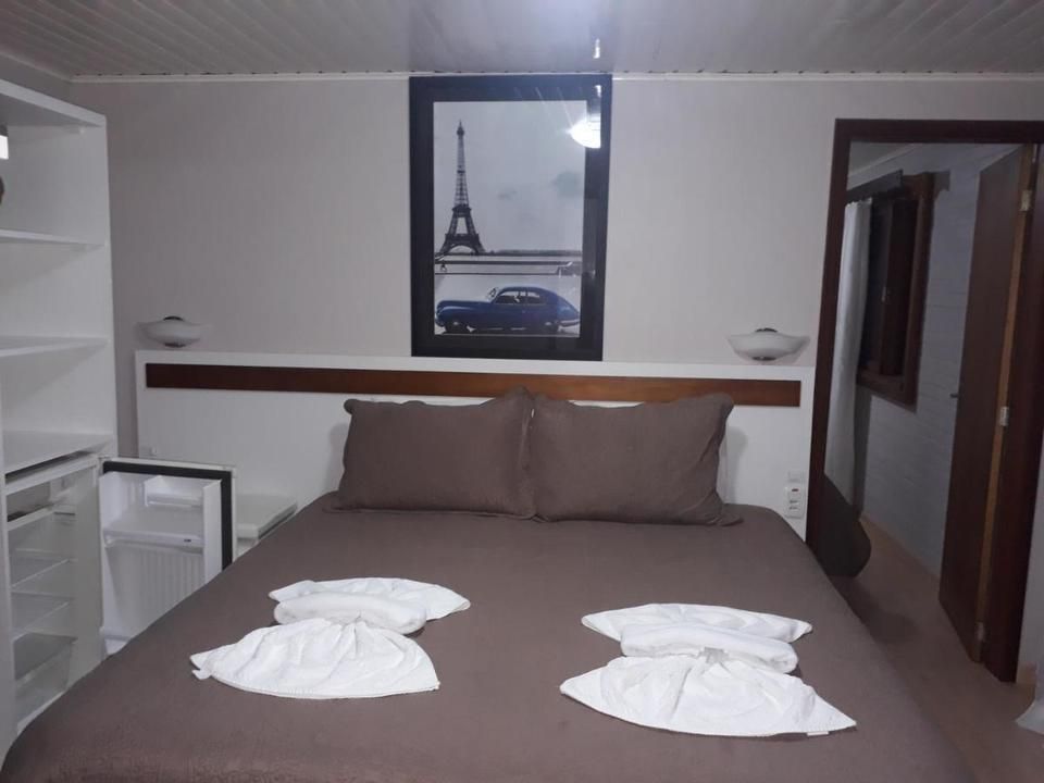 Interior do quarto com cama, roupa de cama marrom, toalhas brancas, um quadro da Torre Eiffel e um frigobar.