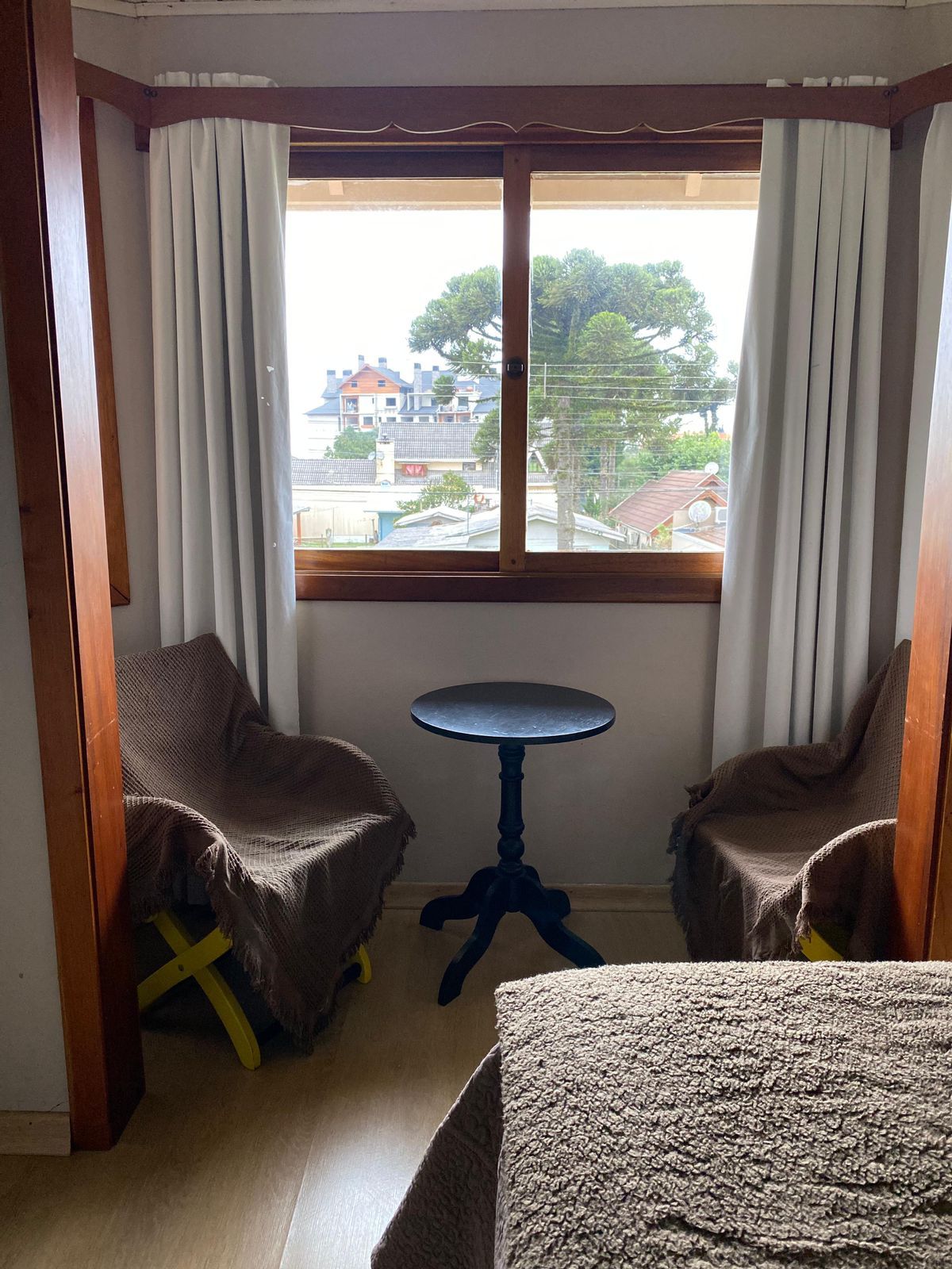 Área de estar aconchegante com duas cadeiras, uma pequena mesa, janela com cortinas e vista para árvores e edifícios.