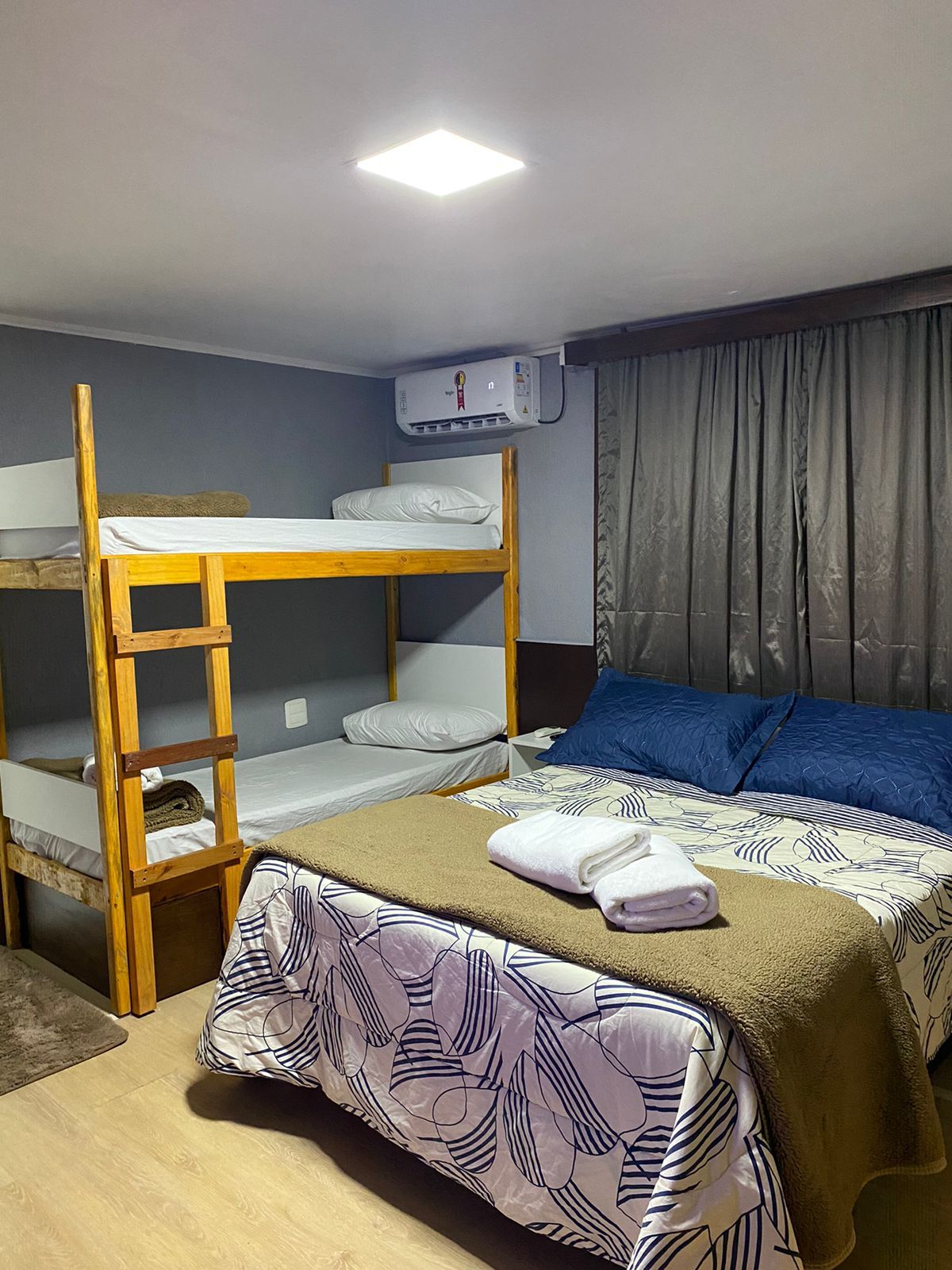 Quarto com beliches, cama de casal e ar condicionado. Paredes cinza, roupa de cama azul e marrom e toalhas brancas.