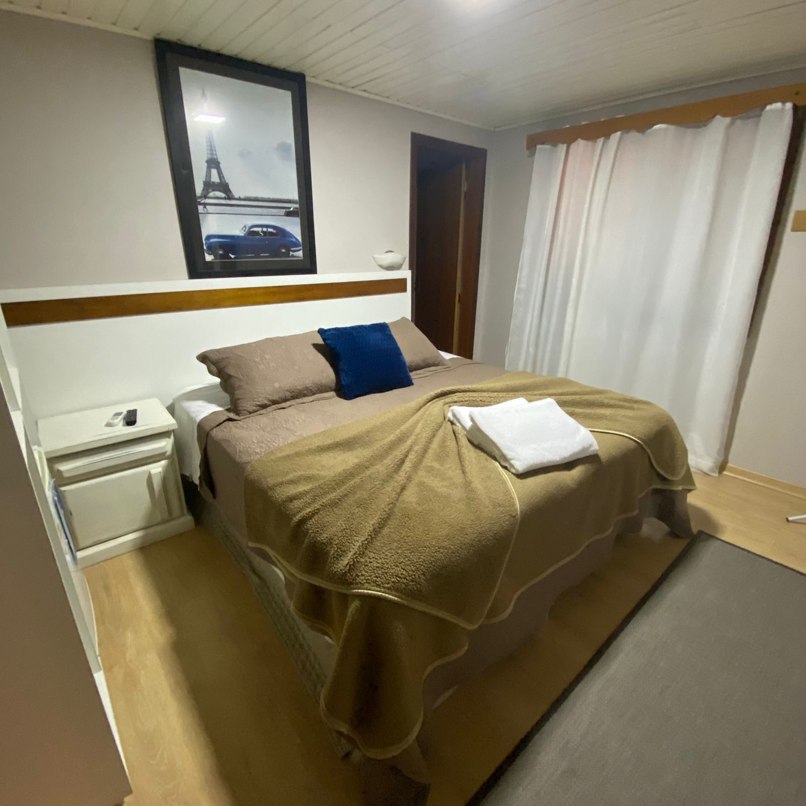 Quarto aconchegante com cama de casal, mesa de cabeceira e quadro da Torre Eiffel. Tons neutros com roupa de cama bege.