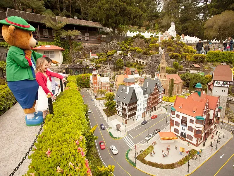 Menina com urso fantasiado em uma vila em miniatura. Prédios coloridos, vegetação e veículos.