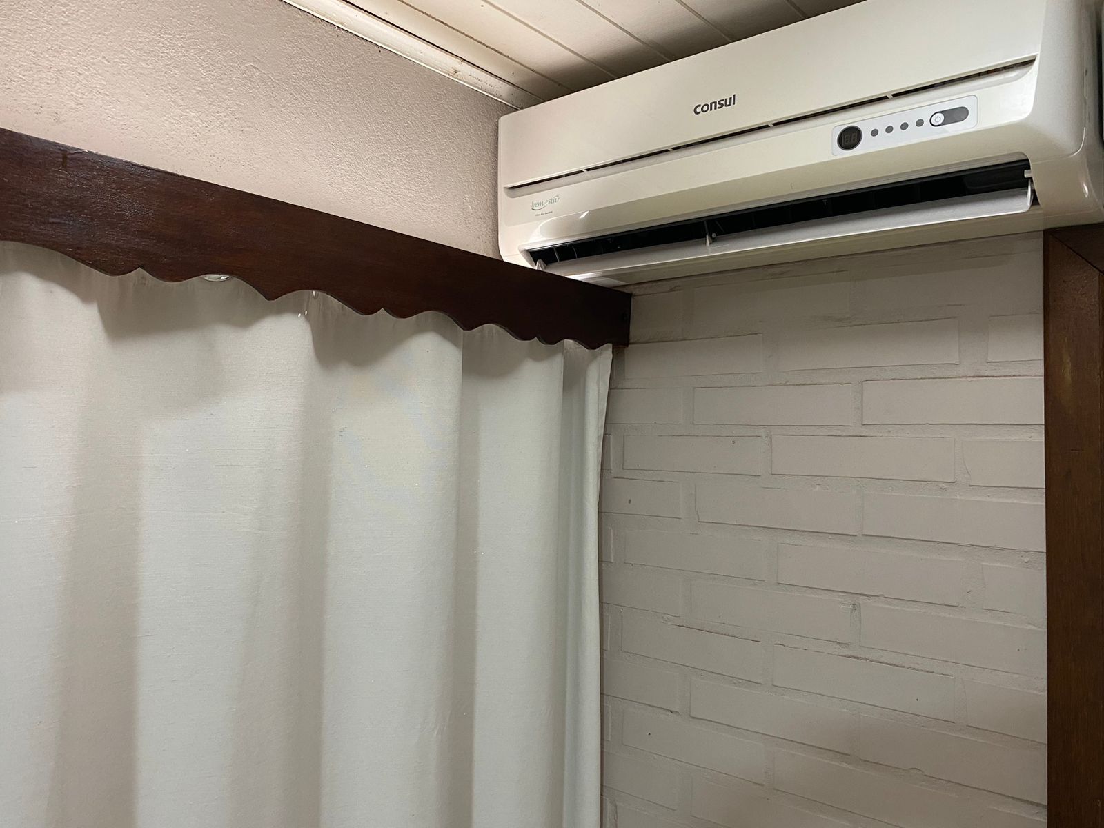 Aparelho de ar condicionado branco instalado acima de uma parede de tijolos brancos e cortina cor creme. Uma moldura de madeira escura está acima da cortina.