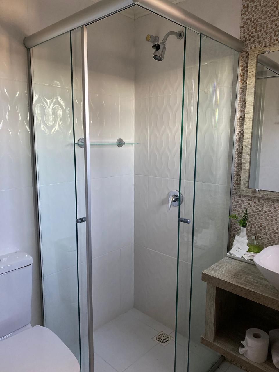 Box de canto com portas de vidro, azulejos brancos e acessórios prateados. O vaso sanitário e a pia também são visíveis.