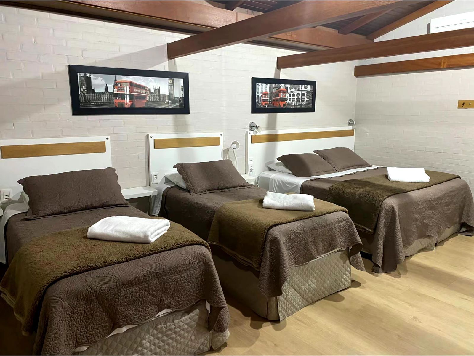Três camas em um quarto, cada uma com roupa de cama marrom e toalhas brancas. Obra de arte com um ônibus londrino acima.