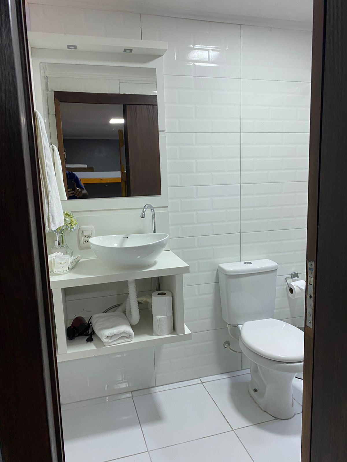 Banheiro com azulejos brancos, pia e vaso sanitário. Um espelho reflete uma beliche.