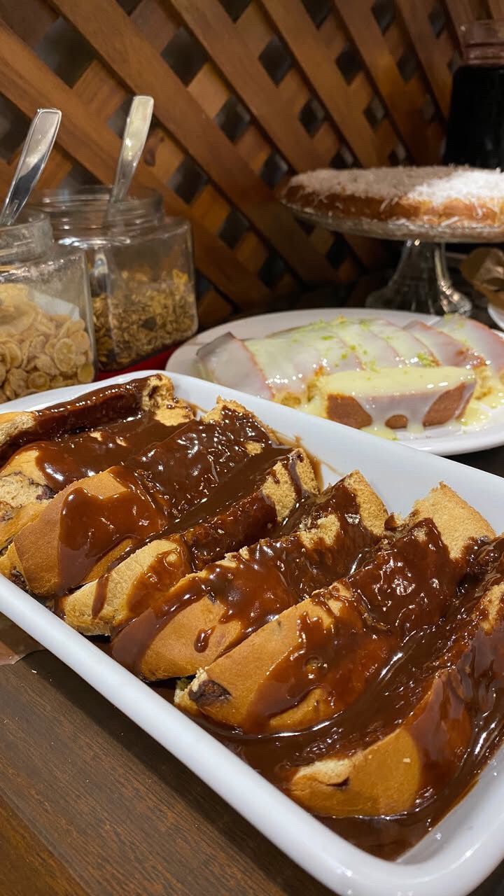 Fatias de doces com calda dispostas em um prato branco, com outras sobremesas e potes de coberturas ao fundo.
