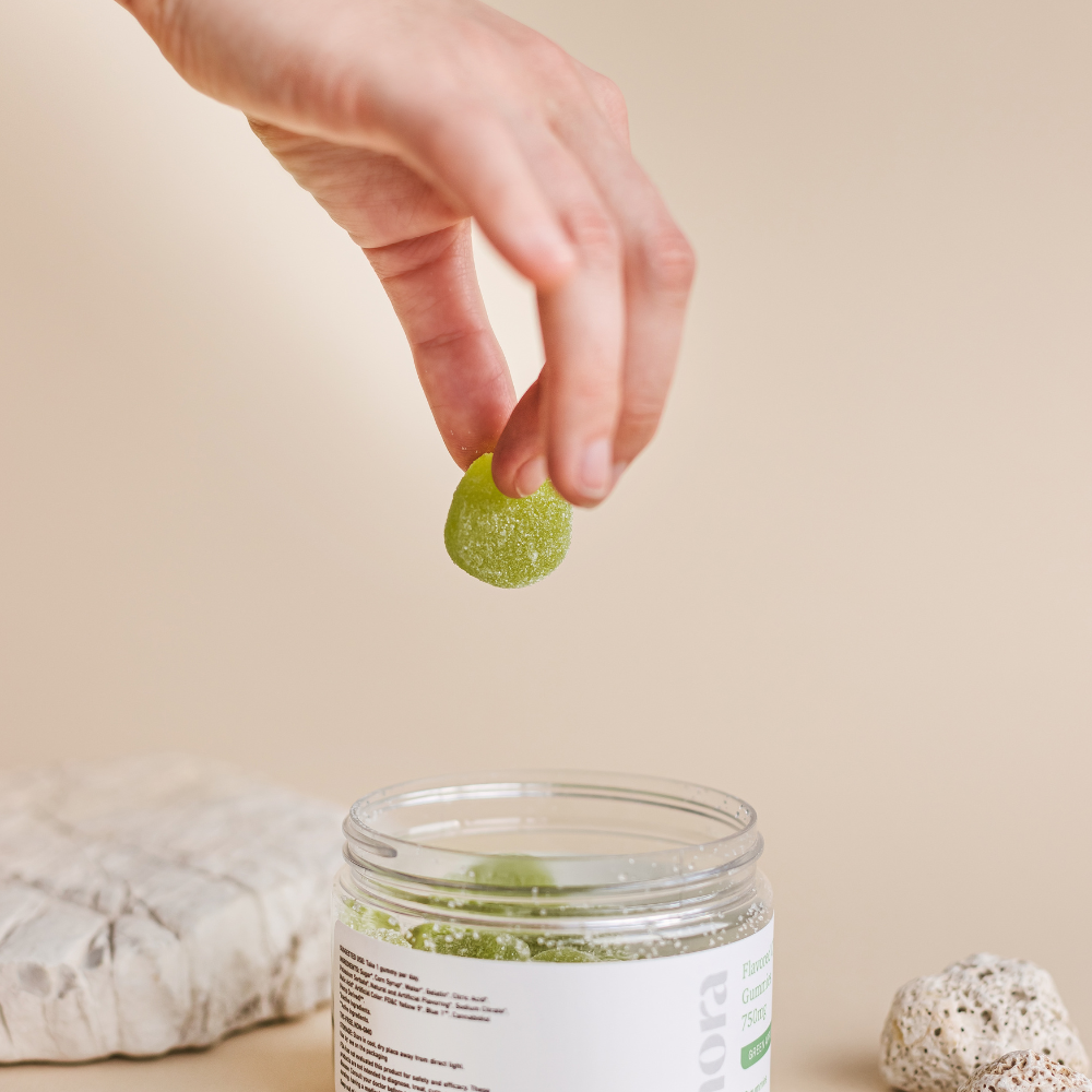 Hand holding a green gummy over a jar. Beige background.