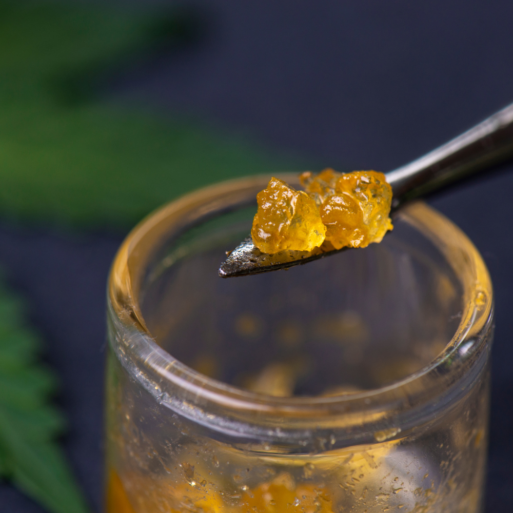 Tweezers holding amber-colored cannabis concentrate above a glass jar, dark background.