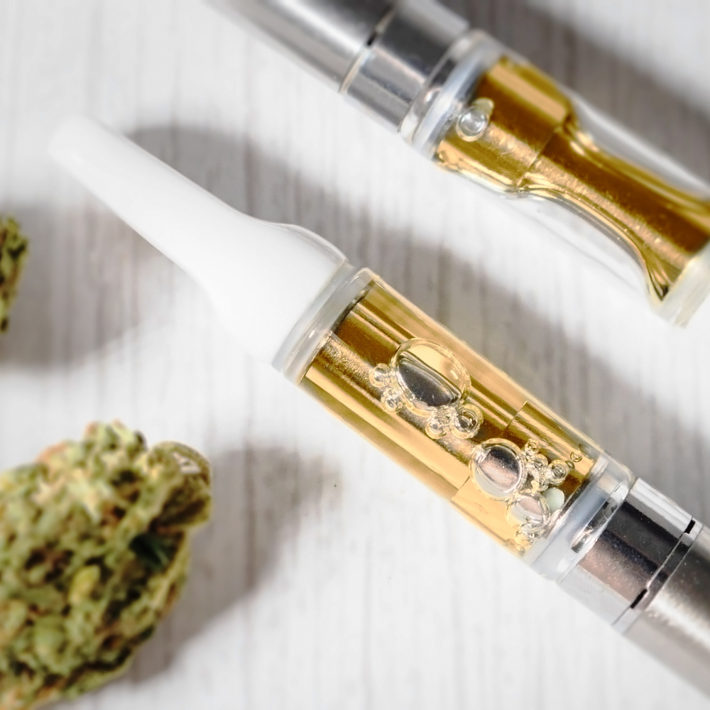 Two cannabis vape cartridges with gold-colored liquid, buds of cannabis on white surface.