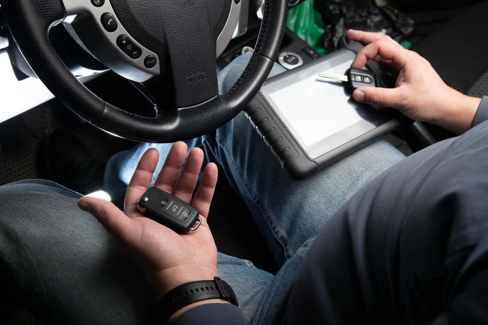 A technician programs a car key fob using a tablet while sitting in the driver's seat of a vehicle.