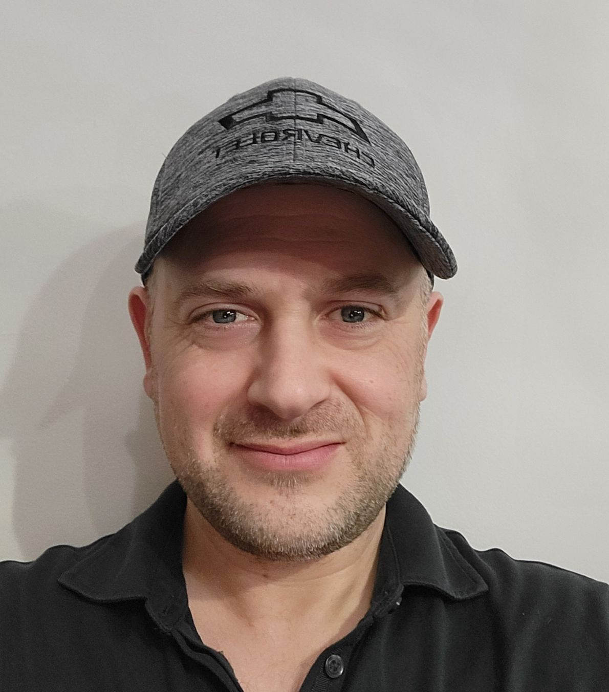 A person wearing a grey Chevrolet baseball cap and a black polo shirt, smiling slightly against a plain, light background.