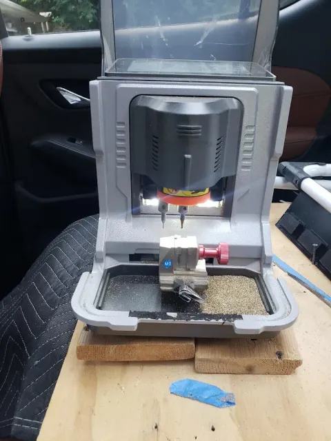A portable electronic key-cutting machine sits on wooden blocks inside a vehicle, with metal shavings in its collection tray.