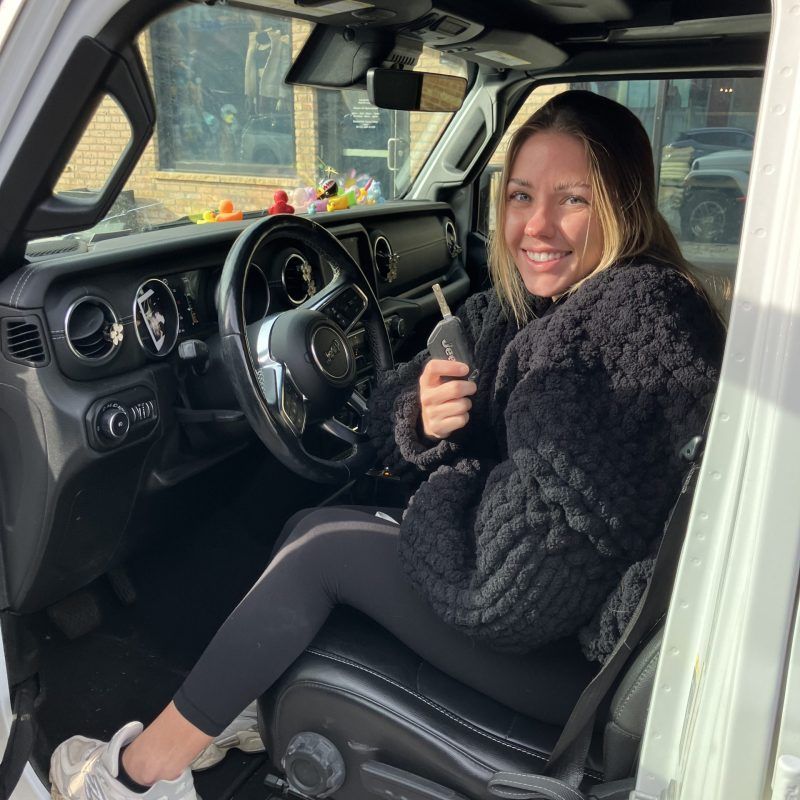 A smiling person sits in the driver's seat of a vehicle, holding a car key fob.