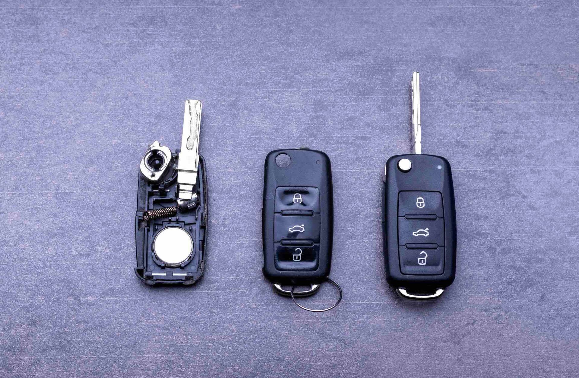 Three car key fobs on a gray surface: one disassembled showing its interior, and two assembled with flip-out keys.