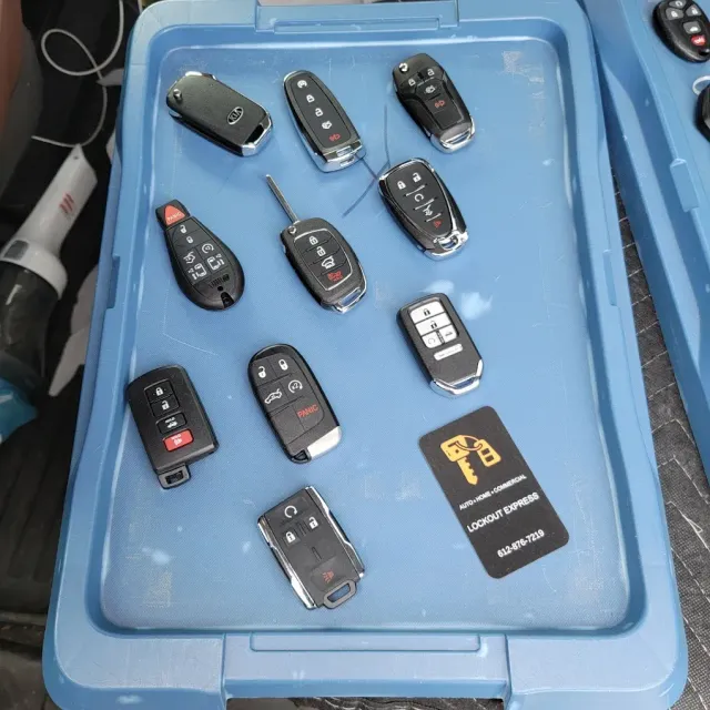 A blue plastic bin containing several assorted car key fobs and a black business card with a locksmith logo.