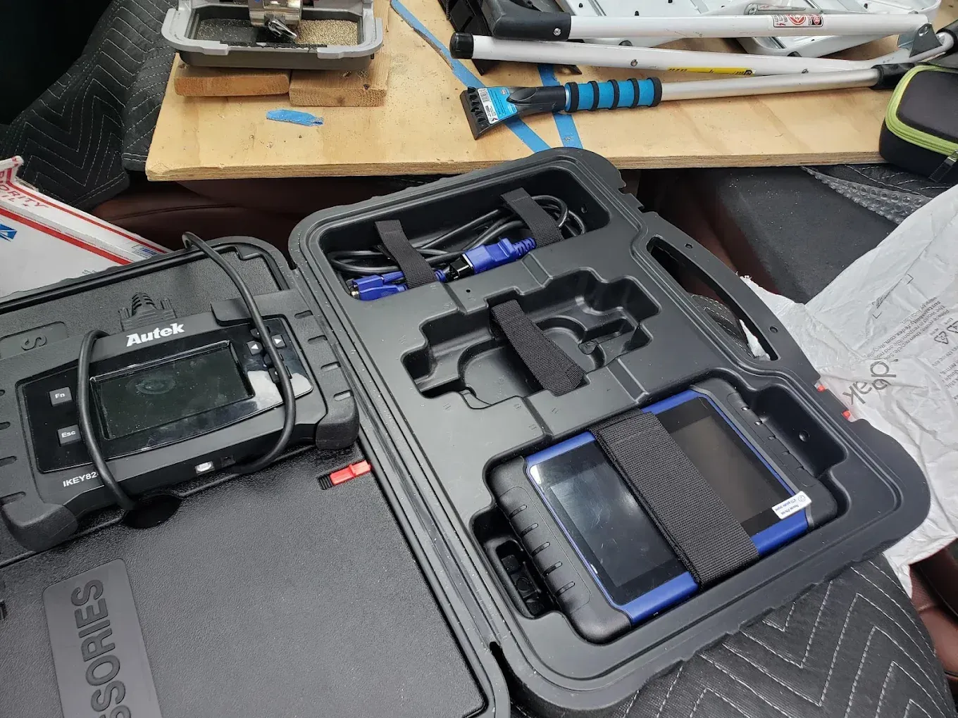 An open case containing two diagnostic scanner tools, with cables and equipment visible on a wooden surface nearby.