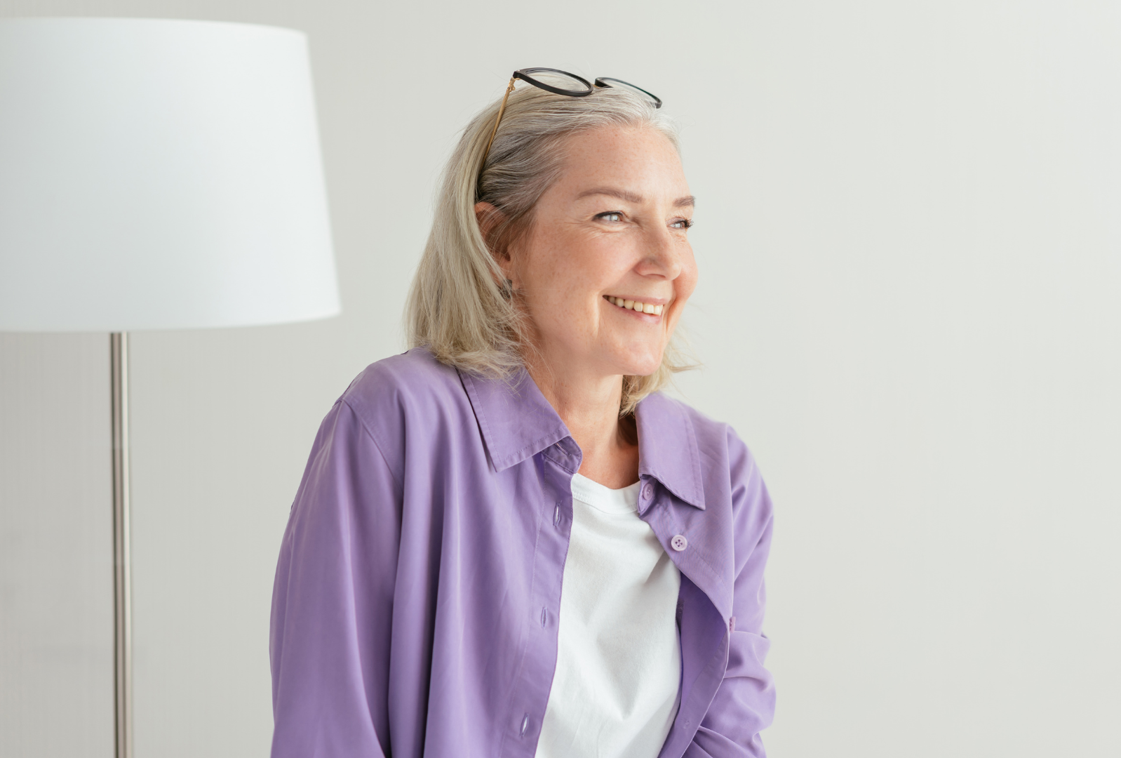 A woman in a purple shirt and glasses is smiling while sitting in front of a lamp.