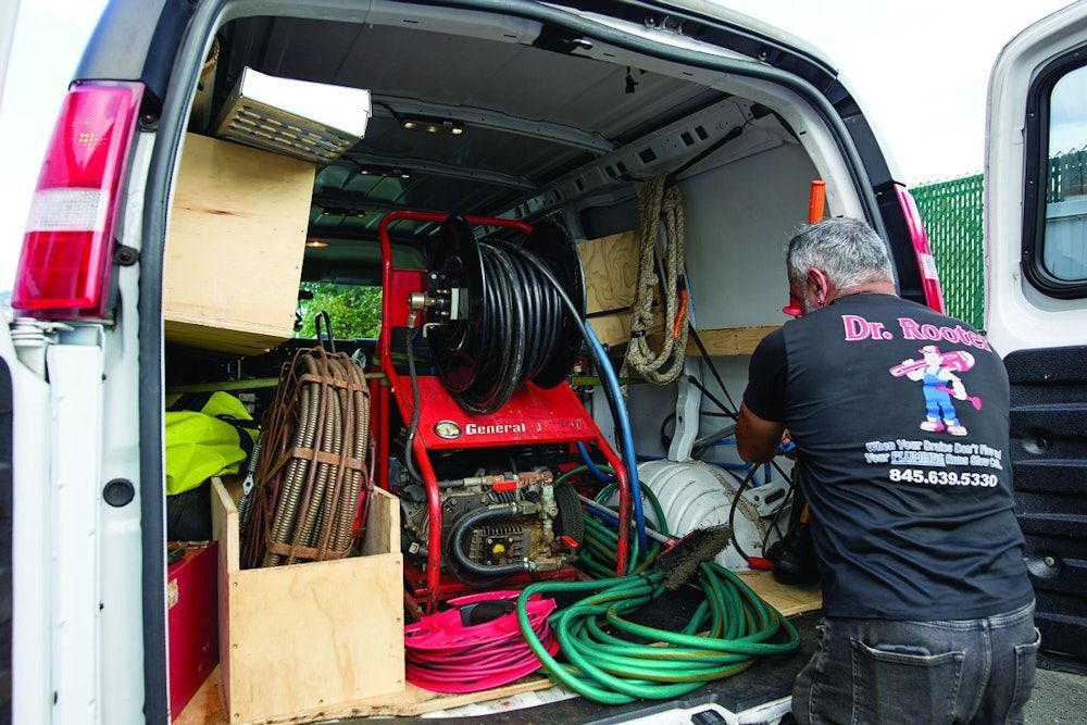 Man in back of van, with tools for drain cleaning. Red machine, hoses, and wood storage.