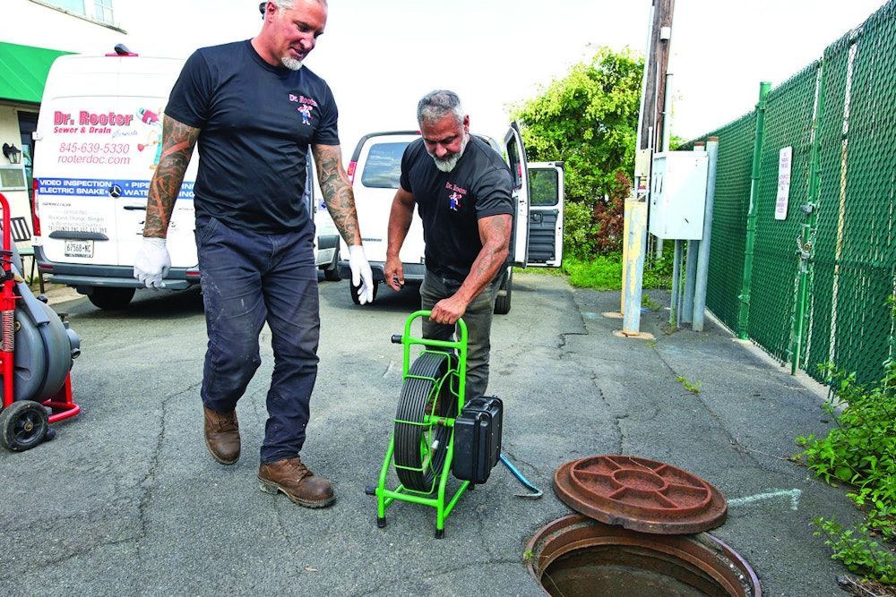 Two men in work attire prepare to inspect a sewer, using a camera on a green cart.