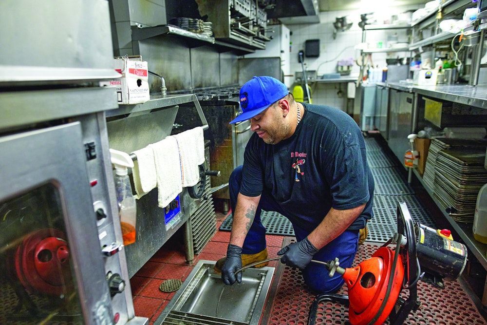 A man in a blue cap kneels in a restaurant kitchen, unclogging a sink with a red drain snake.