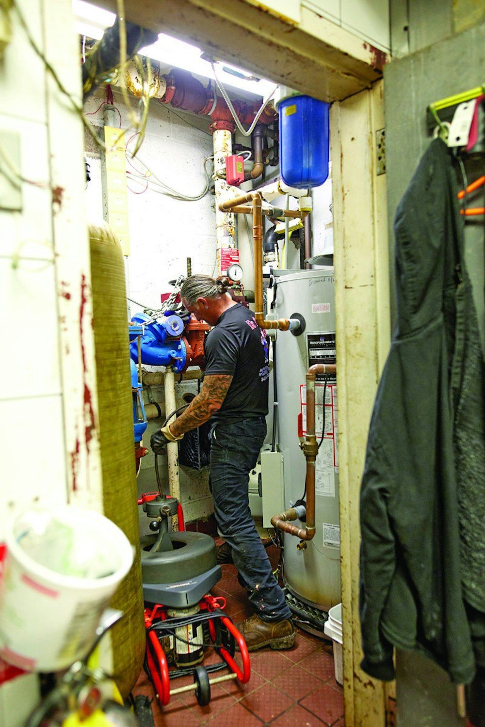 A man works on pipes in a small, cluttered utility room. He's wearing a black shirt and jeans.