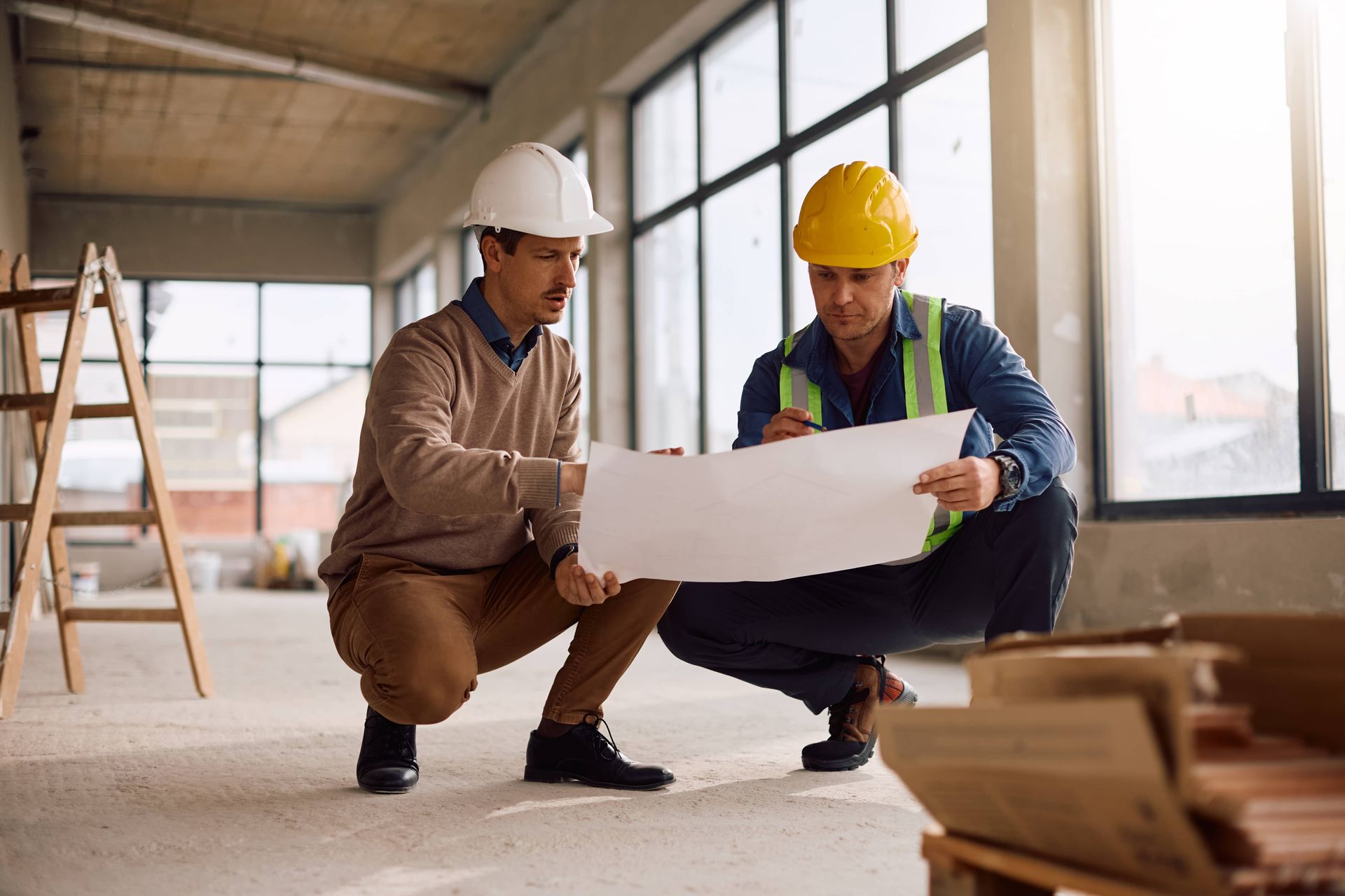 Two construction workers in hard hats reviewing blueprints inside a building under construction.