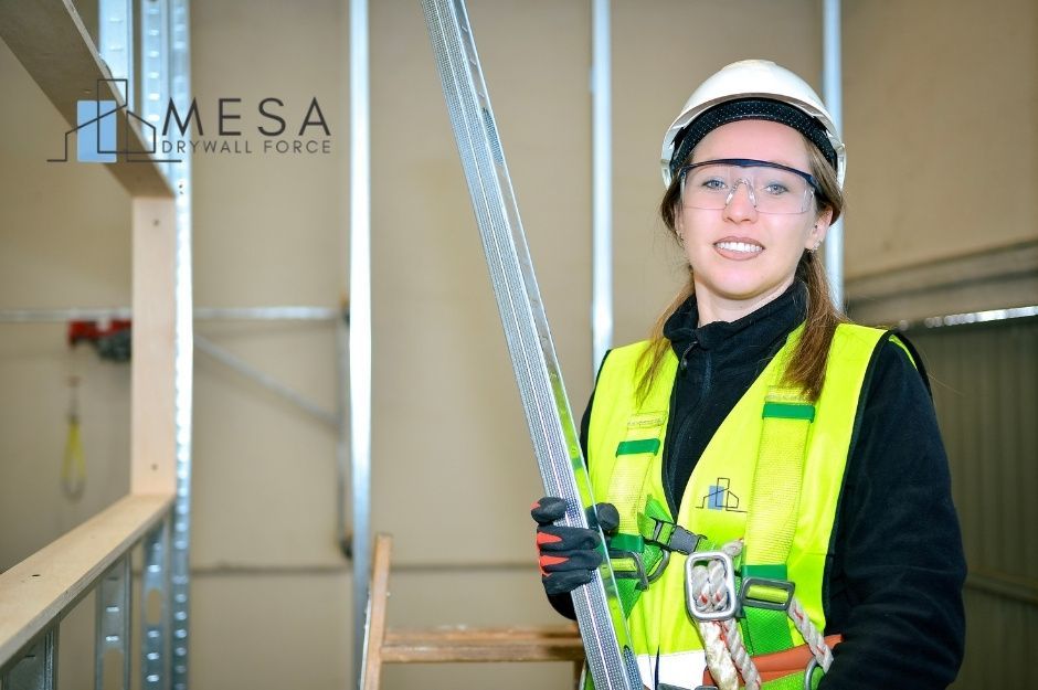 A drywall repair company worker wearing a yellow high-visibility safety vest and white hard hat smiles while holding a metal drywall framing stud at a residential construction site with drywall panels visible near E Red Dog Dr, and N Canyon Ridge Trail, Cave Creek, AZ.