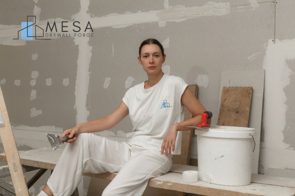A drywall repair company worker wearing a white outfit sits on a wooden plank, holding a putty knife beside a white bucket in an unfinished commercial room with patched drywall and tools scattered around near N Grand Canyon Dr, and W Pueblo Dr, Eloy, AZ.