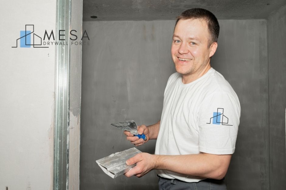 A drywall repair company worker smiles while holding a putty knife and hawk during a commercial project. He wears a white shirt and stands near freshly applied drywall compound on the wall near E Calle Magdalena, and Cll Sahuaro, Guadalupe, AZ.