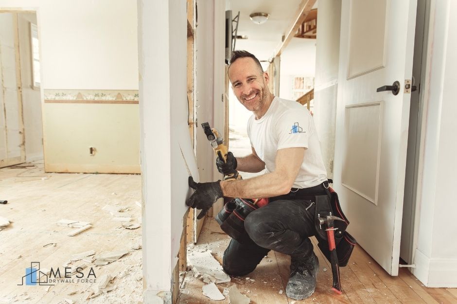 A drywall repair contractor is kneeling in a residential construction site hallway, smiling while holding an oscillating multi-tool. He wears a white t-shirt, dark pants, work gloves, and a tool belt. Debris covers the floor around him near E Summit View Dr, and E Ridgestone Dr, Fountain Hills, AZ.