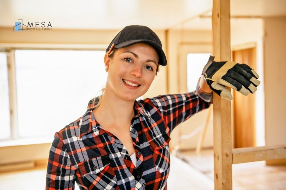 A drywall repair contractor wearing a plaid shirt, black cap, and work gloves is standing in a residential construction area with exposed wooden framing and drywall panels visible in the background near N 70th Ln, and W Peak View Rd in Peoria, AZ.