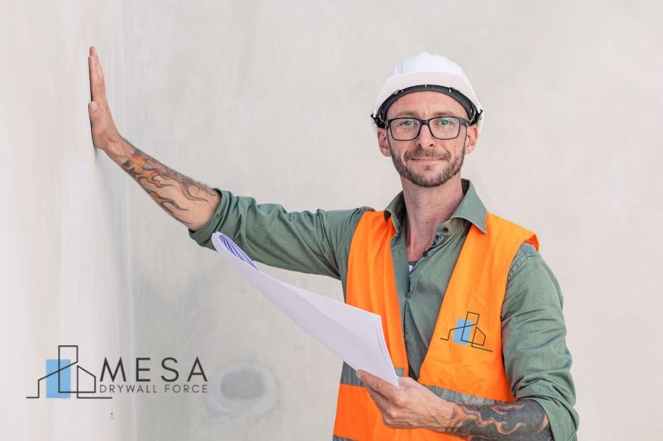 A drywall repair contractor, wearing an orange safety vest, green shirt, and white hard hat, holds a commercial construction blueprint in one hand while leaning toward a wall with the other. He is inspecting work near Echo Canyon Rd, and W Twin Peaks Pkwy, San Tan Valley, AZ.
