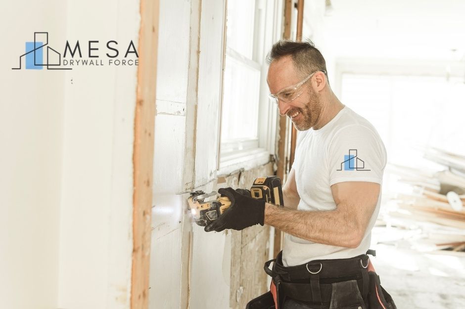A drywall repair contractor wearing safety glasses and gloves uses a cordless oscillating multi-tool to cut into a section of drywall inside a bright residential home under renovation. Exposed framing and stacked materials are visible in the background. Located near N 103rd St, and N 103rd Way, Scottsdale, AZ.