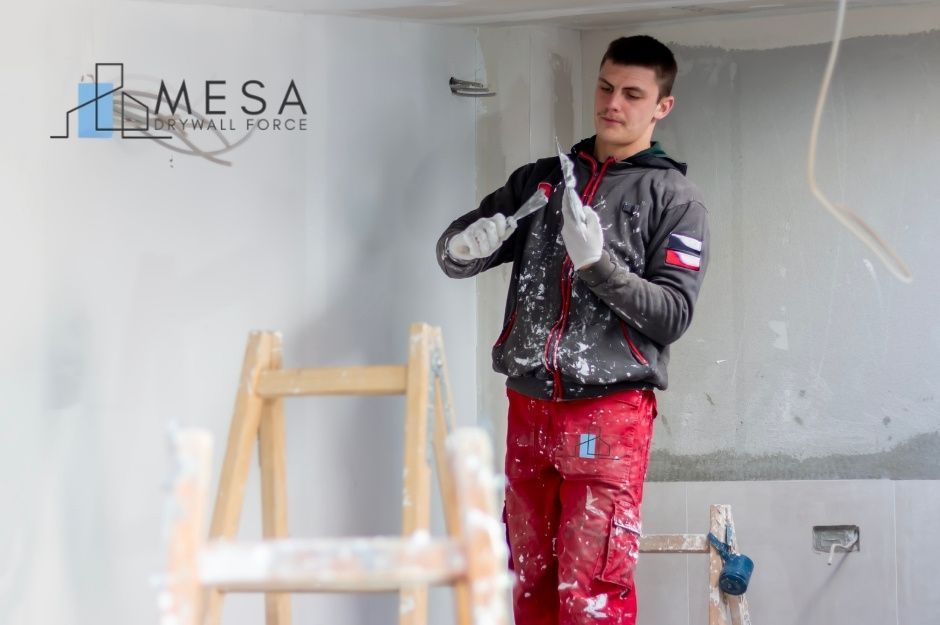 A drywall repair contractor wearing paint-splattered red work pants and a dark gray jacket smooths joint compound using two drywall knives in a residential room under renovation, with a wooden ladder and unfinished white walls visible near N 86th Ln, and W Monroe St, Tolleson, AZ.